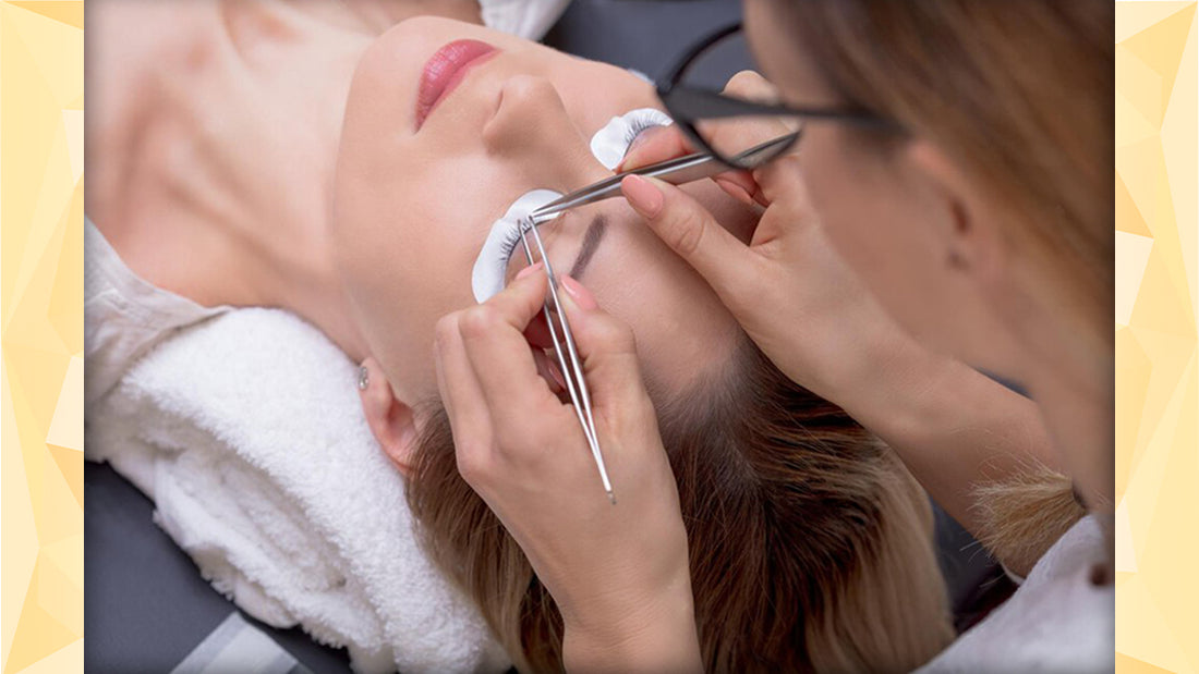 Image of a lash artist holding two tweezers while putting on an eyelash extension to a client who is lying on a white towel in a lash bed.