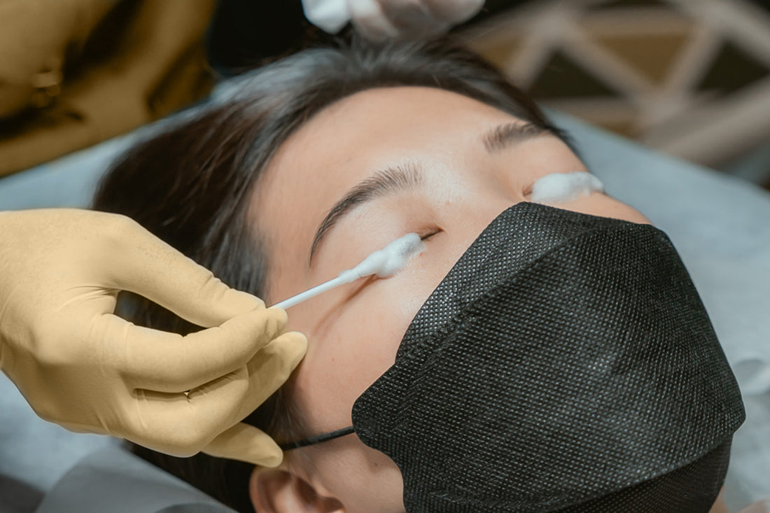 An image of a woman with a black face mask with someone shampooing here eyelashes