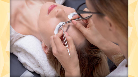Image of a lash artist holding two tweezers while putting on an eyelash extension to a client who is lying on a white towel in a lash bed.